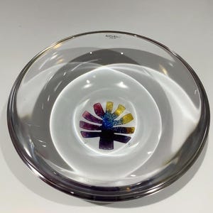 Könnte beinhalten: Eine klare Glasschale mit einem glatten, kreisförmigen Rand und einer tiefen Mulde. In der Mitte des Bodens befindet sich eine stilisierte, mehrfarbige Grafik, die an einen Baum oder ein Blatt in Violett-, Blau- und Gelbtönen erinnert. Der Text KOSTA BODA ist auf dem inneren Rand aufgedruckt.