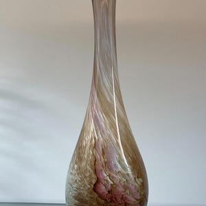 Puede incluir: Un jarrón de vidrio alto y esbelto con una base en forma de lágrima y un cuello estrecho. El vidrio presenta un patrón de remolinos en tonos marrones, blancos y rosa suave translúcidos, creando un efecto jaspeado en la superficie lisa.
