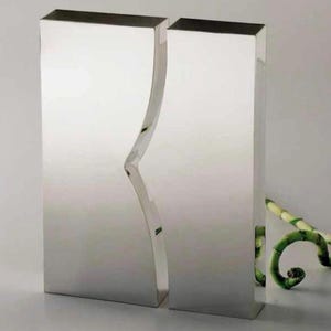Puede incluir: Dos jarrones rectangulares espejados de diseño moderno. Un jarrón tiene una hendidura curva en forma de ola. Los jarrones son plateados y reflejan el entorno. Tallos de bambú verdes se colocan cerca de los jarrones.