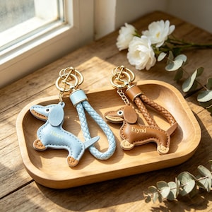 May include: Two dachshund keyrings with gold-coloured hardware. One is light blue with a braided strap, the other is brown with a braided strap. Both are in a wooden tray with white flowers.