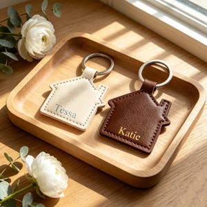 Puede incluir: Dos llaveros con forma de casa y anillas metálicas. Uno es de color crema con el nombre Tessa grabado en gris, y el otro es de color marrón oscuro con el nombre Katie grabado en dorado. Ambos tienen costuras visibles en los bordes.