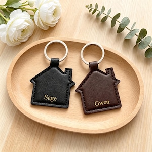 May include: Two house-shaped keychains with silver metal rings. One keychain is black with the name Sage embossed in gold, and the other is dark brown with the name Gwen embossed in gold. Both feature stitched edges and a smooth, flat surface.
