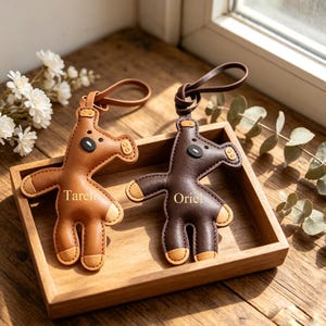May include: Two leather horse-shaped keyrings in a wooden tray. One is light brown with the name "Taren" and the other is dark brown with the name "Orie". Both have tan accents and a loop for attaching keys.