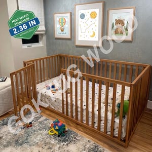 Könnte beinhalten: Ein helles Holzbodenbett mit vertikalen Latten und einem Seitengitter. Das Bett hat eine weiße Matratze mit dunklem Muster und ein grünes Sicherheitsetikett mit der Angabe 6 cm zwischen den Stäben. Drei gerahmte Wandbilder hängen über dem Bett.