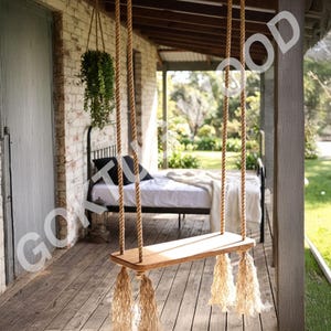 Op de afbeelding: Een houten schommel met touwsteunen en decoratieve kwasten hangt aan een veranda. De zitting van de schommel is lichtbruin en de touwen zijn naturel beige. De achtergrond toont een bed en een bakstenen muur.