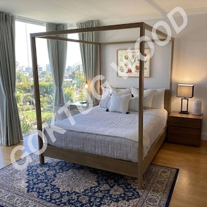 May include: A light brown wooden canopy bed frame with a light grey textured quilt and several white pillows. The bed is positioned in a room with light grey curtains, a blue and cream patterned area rug, and two wooden nightstands with lamps.