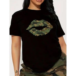 May include: Black short-sleeved t-shirt featuring a camouflage lips graphic. The lips are in shades of green, brown, and tan. The shirt is paired with camouflage pants.