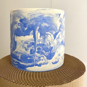 Pode incluir: Um vaso cilíndrico com um design marmoreado em tons de azul e creme. O vaso está sobre um suporte de cartão castanho com padrão de favo de mel. É um artigo decorativo para plantas de interior.