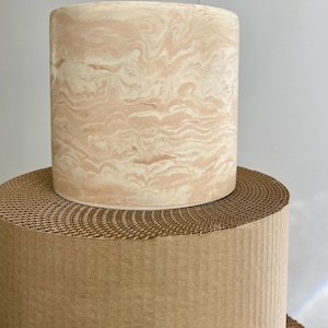 Pode incluir: Um recipiente cilíndrico de cerâmica com um padrão em espiral de pêssego e creme repousa sobre um rolo de cartão ondulado. O design do recipiente assemelha-se ao mármore, adicionando um toque de elegância.