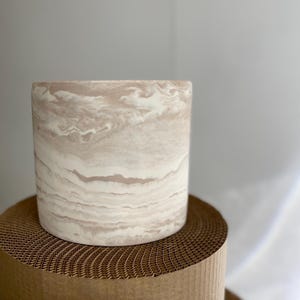 May include: A cylindrical planter with a marbled beige and cream pattern. The planter sits on a brown, honeycomb-patterned cardboard roll. The neutral tones and organic design suggest a modern aesthetic.