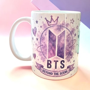 Könnte beinhalten: Ein weißer Keramikbecher mit einer violetten Aquarellgrafik des BTS-Logos, einer Krone, Sternen und Herzen. Der Text "BTS" und "BEYOND THE SCENE" ist in Dunkelviolett auf einem Banner am unteren Rand des kreisförmigen Designs gedruckt.