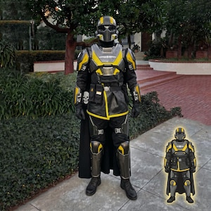 Könnte beinhalten: Ein Ganzkörperkostüm in Schwarz und Gelb, bestehend aus Helm, Rüstung und Umhang. Das Design beinhaltet metallische Akzente und ein Totenkopf-Emblem. Die Gesamtästhetik deutet auf ein futuristisches oder Science-Fiction-Thema hin.