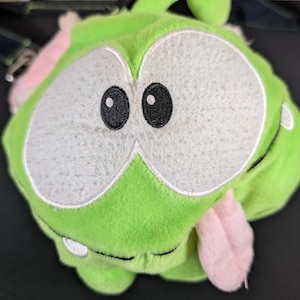 以下が含まれることがあります： 大きな白い目と黒い瞳を持つ、緑色の丸いキャラクターの形をしたぬいぐるみ。キャラクターはピンクの舌を出しており、上部に緑色の茎があります。おもちゃは柔らかいぬいぐるみ素材で作られています。