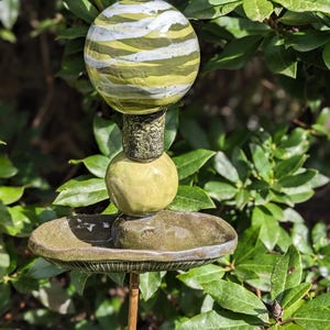Könnte beinhalten: Ein dekorativer Gartenstecker mit einer geschichteten, gestreiften Kugel in Grün-, Weiß- und Gelbtönen. Die Kugel sitzt auf einem Stapel keramischer Elemente, darunter eine kleine Kugel und eine flache Schale, alles in Erdtönen.