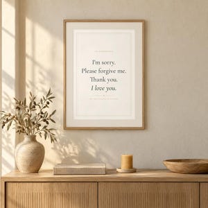 Puede incluir: Una lámina de arte enmarcada con el texto: I'm sorry. Please forgive me. Thank you. I love you. La impresión está en un marco de madera clara y se muestra en una pared sobre una superficie de madera con un jarrón, un libro, una vela y un cuenco.