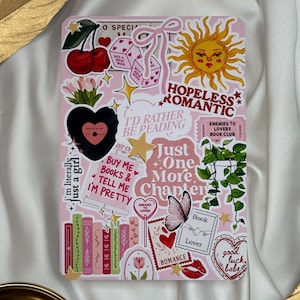 Può includere: Un quaderno rosa decorato con adesivi a tema romantico. Gli adesivi includono ciliegie, dadi, un sole, cuori, libri e frasi come "Hopeless Romantic" e "Just One More Chapter."