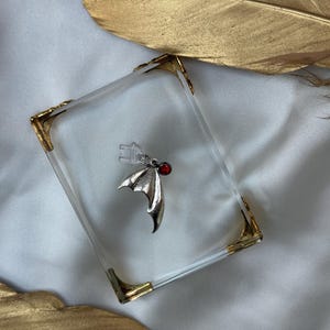 Peut inclure: Un pendentif en forme d'aile de chauve-souris argenté orné d'une petite pierre précieuse ronde rouge foncé sur le dessus. Le bijou comprend un fermoir en plastique transparent fixé à l'anneau, avec un design texturé et une pointe incurvée.