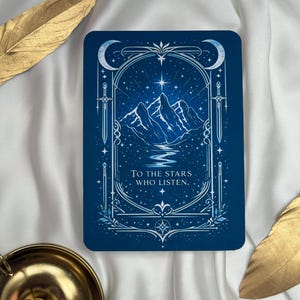 Può includere: Carta rettangolare con sfondo blu scuro raffigurante una catena montuosa sotto un cielo stellato. La carta ha un bordo decorativo con lune crescenti e spade. Il testo "TO THE STARS WHO LISTEN" è visualizzato.
