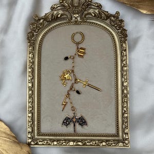 May include: A gold-toned hoop earring featuring a dangling chain with multiple charms including an open book, a dragon, a sword, a lightning bolt, two small black oval beads, and a large black and gold bat-shaped pendant with a central star detail.