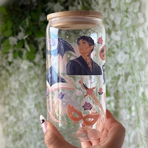 Op de afbeelding: Een doorzichtig glas met een bamboe deksel voorzien van een print van een mannelijk figuur met donker haar, een vleermuisvleugel, een open boek, pijl en boog, een zwaard en een oranje masker, versierd met kleine roze bloemen en sterren.