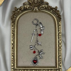 May include: A silver earring with various charms: a sword, bird, star, and a leopard. It also has red crystal accents and a heart pendant. The earring is presented within a gold-framed picture.