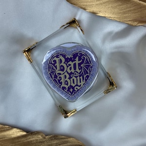 May include: A heart-shaped pin featuring a purple background with a dark purple spiderweb pattern and the text Bat Boy in a decorative font. The pin is encased in a clear square acrylic frame with gold-toned metal corner accents.