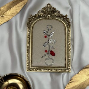 Op de afbeelding: Een zilverkleurige bedel-sleutelhanger met een verzameling bungelende metalen hangers, waaronder een revolver, een schedel, een zwaard, een hart en speelkaarten, geaccentueerd met kleine rode kralen en een ronde ring aan de bovenzijde.