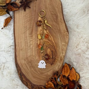 May include: A gold-toned metal keyring featuring a white ghost charm, a small gold horn, a star, and several autumn-themed charms including orange pumpkins and brown maple leaves attached to a circular ring with a lobster clasp closure.