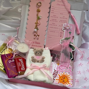 Könnte beinhalten: Eine Geschenkbox mit einem goldfarbenen Anhänger mit Blumen- und Schleifenmotiven, einer rosa Buchbewertungskarte, einem Lotus-Lesezeichen, weißen Socken mit Teddybär-Motiv, einem Biscoff-Keks, einem Pukka-Teebeutel und einer kleinen Kerze.