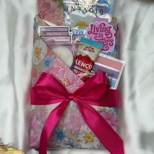Puede incluir: Un sobre de regalo envuelto en papel floral con un lazo de cinta de satén rosa brillante. El sobre contiene una mascarilla facial Biovene, un sobre de café helado Kenco, una vela de té blanca y varias pegatinas, incluido el texto Living in Chapters.
