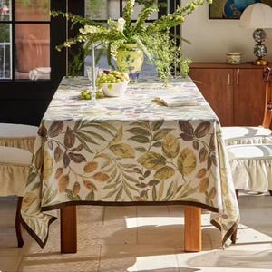 May include: A rectangular tablecloth featuring a botanical print of green, yellow, and brown leaves on a cream background with a solid dark brown border. The fabric drapes over a wooden table, which is set with a white bowl of green grapes.