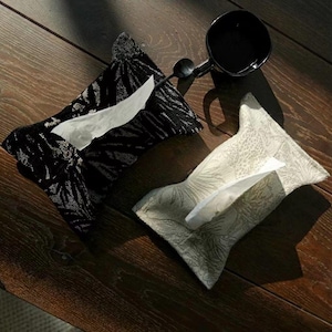 Può includere: Due copri scatole di fazzoletti decorativi su una superficie di legno. Uno è nero con un motivo a foglie argentate, l'altro è bianco sporco con un design floreale. I fazzoletti bianchi sono visibili in ogni scatola.