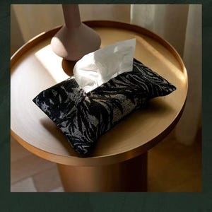 May include: A black and white patterned tissue box cover with white tissues peeking out. The cover sits on a round, gold-toned table. A light pink vase is also on the table. The scene is well-lit, suggesting an interior setting.
