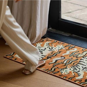 May include: A rectangular rug with a tiger design in orange, black, and white. The rug features two tigers and abstract wave patterns. The rug is placed near a door and a person's leg and foot are visible.