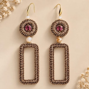 Puede incluir: Un par de pendientes colgantes con una parte superior circular de ganchillo color bronce con un cristal magenta central, una pequeña cuenta blanca y una gran base rectangular de ganchillo color bronce. Ganchos de metal dorado.