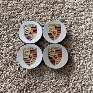 May include: Four circular silver-coloured wheel centre caps, each featuring the Porsche crest in the centre. The crest displays the word PORSCHE at the top, with black and red horizontal stripes, gold background details, and a black horse emblem.