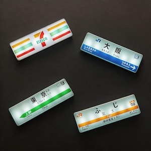 以下が含まれることがあります： 4つの光る長方形の看板。1つはセブンイレブンのロゴ。その他は日本語と英語の翻訳が表示され、「東京」、「大阪」、「富士」など。各看板は光る効果があります。