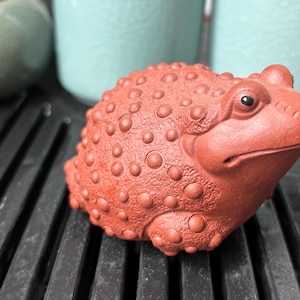 May include: A terracotta-colored ceramic toad figurine featuring a textured back covered in raised, circular bumps. The toad has a wide mouth, defined eyes, and a matte finish, crafted in a squat, rounded shape with a detailed, bumpy skin texture.