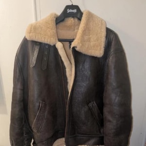 May include: A brown leather bomber jacket with a cream-colored shearling collar and lining. The jacket has a zip-up front, two front pockets, and a shearling trim on the cuffs and hem. The jacket is hanging on a black hanger.