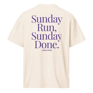 Puede incluir: Camiseta color crema con el texto morado "Sunday Run, Sunday Done." y el nombre de la marca "cotton cartel". La camiseta tiene mangas cortas y un corte relajado. El texto está en una fuente elegante.