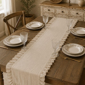 Puede incluir: Una mesa de comedor rústica preparada para una comida. La mesa tiene un camino de mesa beige con volantes. La mesa está puesta con platos, vasos y cubiertos. Los platos tienen un diseño floral.