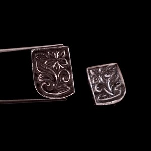 May include: Two clear, shield-shaped gemstones featuring an intricate, carved floral pattern with petals and swirling leaves. One gemstone is held by a pair of metal tweezers, while the other rests nearby, both displaying a polished, translucent surface.