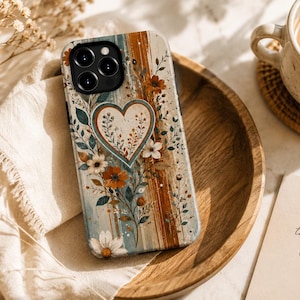 Puede incluir: Una funda para smartphone con un corazón central rodeado de ilustraciones botánicas de flores y hojas en tonos naranja óxido, azul cerceta y crema. El fondo tiene una apariencia texturizada con trazos de pincel verticales de color.