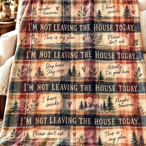 May include: A plaid throw blanket in shades of red, blue, and cream featuring the repeated text I'M NOT LEAVING THE HOUSE TODAY. Additional phrases include maybe tomorrow, I have snacks, This is my plan, Please don't ask, Stay home, Stay cosy, and Actually, I'm good here, accented with small pine tree and heart illustrations.