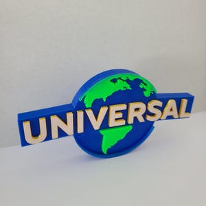 May include: A 3D-printed Universal Studios logo. It features a blue globe with green continents and the word "UNIVERSAL" in beige block letters. The logo is set against a white background.