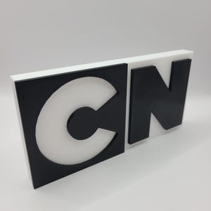 May include: A 3D-printed sign featuring the letters "CN" in a bold, blocky font. The letters are black, set against a white background. The sign has a rectangular shape and is likely a logo or decorative piece.