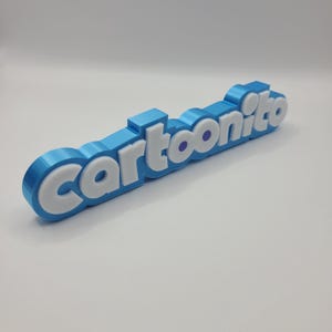 May include: A 3D-printed sign spelling "Cartoonito". The sign is light blue with white lettering and purple accents on the letter "o". The sign is on a white surface.