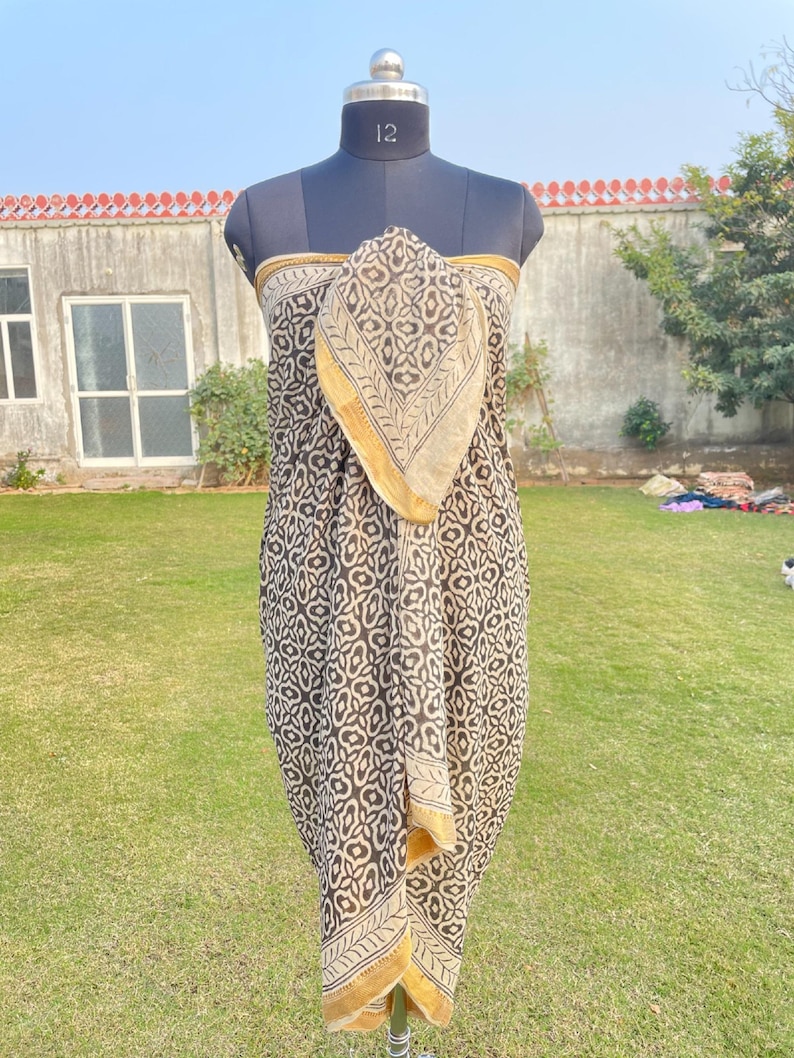 May include: A sarong featuring a dark brown and cream repeating floral pattern with a solid gold border. The fabric is draped over a mannequin torso, showcasing the intricate block-print style design and the contrasting hemline detail.