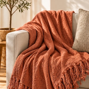 Op de afbeelding: Een terracotta-kleurige plaid met een textuurrijke, geweven stofafwerking en decoratieve kwastjes langs de onderrand. De deken is gedrapeerd over de armleuning en zitting van een lichtgekleurde bank, wat de zachte, gekreukte uitstraling benadrukt.