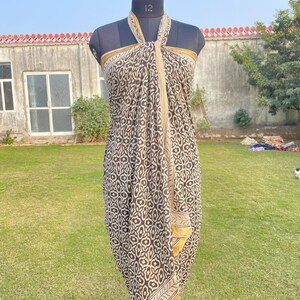 May include: A sarong featuring a dark brown and cream geometric pattern with a repeating floral-like motif. The fabric is draped around a mannequin in a halter style, showcasing a solid cream border along the edges and a soft, flowing silhouette.